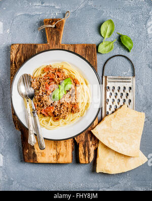 Pasta dinner. Spaghetti Bolognese in metal plate on rustic wooden board with Parmesan cheese grater and fresh basil on grey con Stock Photo