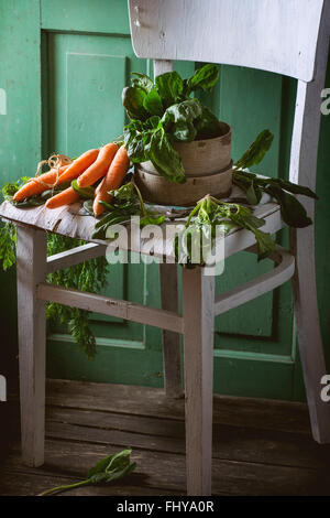 Bunch of fresh spinach and carrots on old white wooden chair with green wooden wall at background.