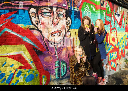 London, UK. 26th February, 2016. Three students enjoying Rainbow beigels, with BrickLane’s street art grafitti in the background. Rainbow bagel. Stock Photo