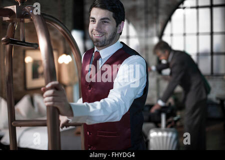Bellboy with trolley, business man with suitcases in background, hotel room Stock Photo