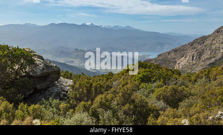 View over St Florent and snow capped mountains in north Corsica with rocks and maquis in the foreground Stock Photo