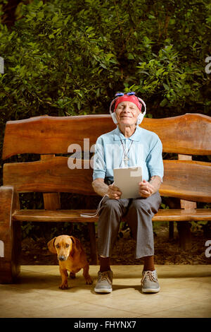 MODEL RELEASED. Senior man sitting on a bench listening to music with his dog. Stock Photo