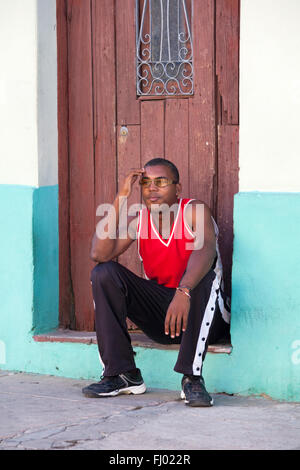 Daily life in Cuba - Cuban young man wearing glasses sitting on doorstep in doorway at Cienfuegos, Cuba, West Indies, Caribbean, Central America Stock Photo