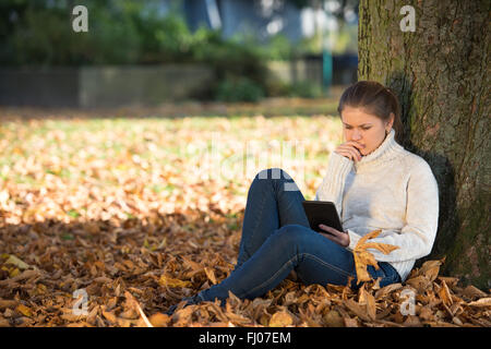 young woman in autumn park sitting near tree reading ebook Stock Photo