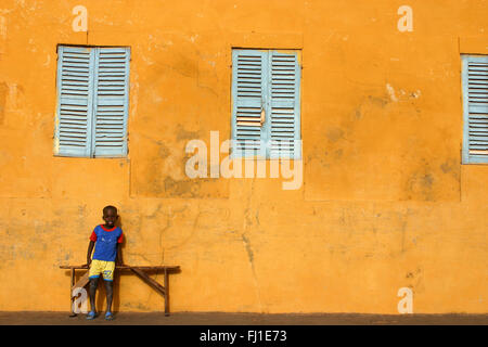 A boy is posing in front of a  wall of a typical colonial house painted in yellow in the streets of Saint Louis, Senegal Stock Photo