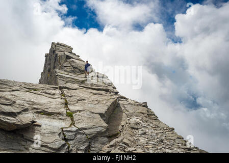 Hiker climbing on steep rocky slope towards the mountain summit without protection. Dramatic cloudy sky. Concept of reaching the Stock Photo