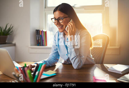 Business woman working on laptop smiling talking on phone sun coming through window Stock Photo