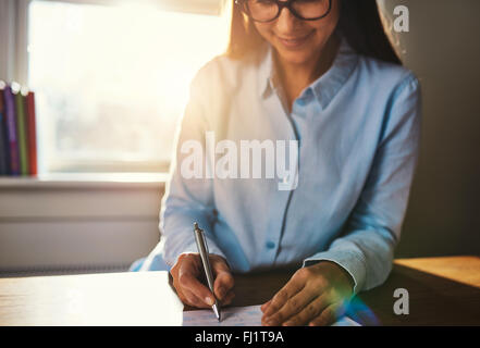Selective focus on hands of smiling woman in blue blouse writing at desk on notepad in home office with sunlight over her should Stock Photo
