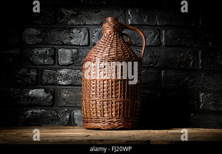 Wicker bottle on wooden table near brick wall Stock Photo