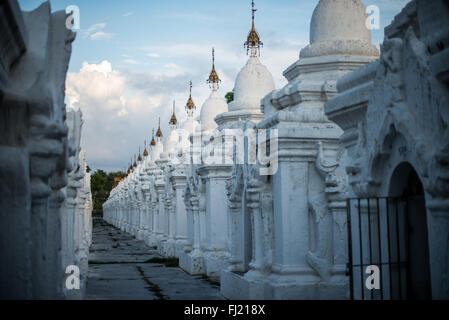 MANDALAY, Myanmar — Rows of pristine white kyauksa gu (stone-inscription caves) stretch across the grounds of Kuthodaw Pagoda. Built in 1857 by King Mindon, the complex contains 729 marble slabs that collectively form what is known as the World's Largest Book. Each small stupa houses a marble tablet inscribed on both sides with text from the Tipitaka, the complete Pali Canon of Theravada Buddhism. The orderly arrangement of these structures reflects traditional Buddhist architectural principles. Stock Photo