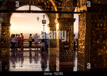 Sutaungpyei Pagoda Sunset Golden Archway Tourists Mandalay Myanmar | MANDALAY, Myanmar — Visitors gather on the terrace of Sutaungpyei Pagoda at sunset, framed here through a mirrored-glass archway whose gold mosaic columns open onto the Ayeyarwady plain at dusk. The summit rises roughly 240 m (790 ft) above the city — high enough that in March 1945 the 19th Indian Division's commander refused an air strike on these very structures to spare the pagodas while his troops fought to clear the hill. Stock Photo