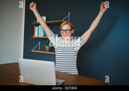 Female entrepreneur at home office with arms raised in success sitting with a laptop
