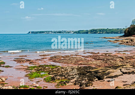 Rocks protruding through the reddish coloured sand contrast strongly with the blue sea, sky and green vegetation on the headland Stock Photo