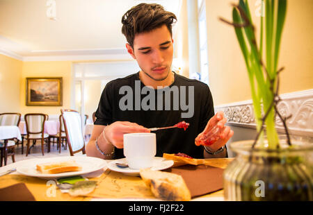Attractive Young Man Eating Breakfast, Having a Slice of Bread with Jam and Butter and Drinking Coffee Stock Photo