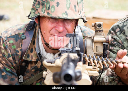 Second World War re-enactment. Waffen SS man in camouflage uniform, behind machine gun with barrel pointing at viewer, eye-contact. Close-up. Stock Photo