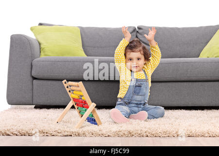 Baby girl sitting on floor in front of a gray sofa and gesturing with her hands with an abacus next to her Stock Photo