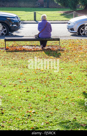 Outdoor seating. Woman sitting outside on a wooden seat near a road on a sunny day in Autumn. Ruddington, Nottinghamshire, England, UK Stock Photo