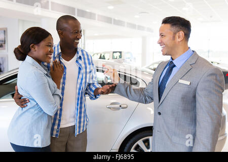 mid age car salesman handing over new car key to African couple in car showroom Stock Photo