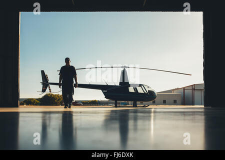 Silhouette of helicopter with a pilot in the airplane hangar. Pilot walking away from helicopter parked outside the hangar. Stock Photo