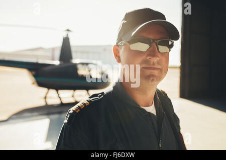 Close up portrait of professional pilot standing in airplane hangar with a helicopter in background on a sunny day. Stock Photo