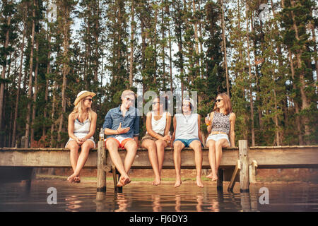 Portrait of young friends sitting on jetty and talking. Young men and women enjoying a day at the lake. Stock Photo