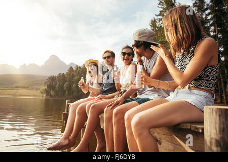 Portrait of happy young friends sitting on pier at the lake drinking beers. Young men and woman enjoying a day at the lake. Stock Photo