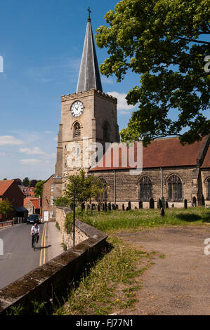 A man cycles on the road, past St Leonard and St Mary Roman Catholic Church, Malton, North Yorkshire, GB - its tall spire set against a blue sky. Stock Photo