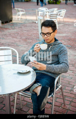 Serious young asian man sitting in relaxing outdoor cafe holding cup of coffee and using mobile phone Stock Photo