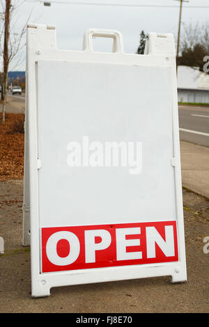 Blank white sign says open place on the sidewalk near a busy street in front of a business. Stock Photo