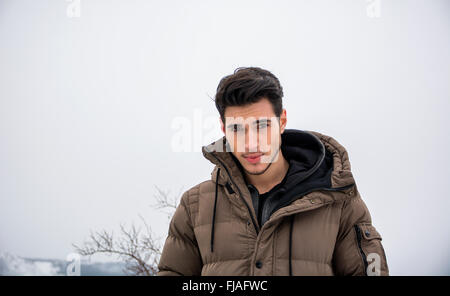 Handsome man in outerwear sitting while looking at camera. Snowy landscape on background Stock Photo