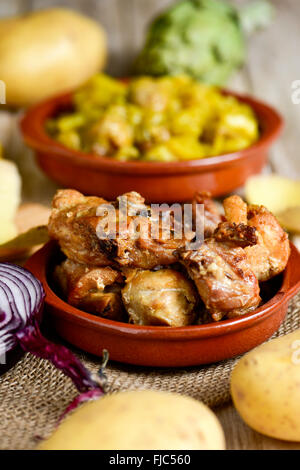an earthenware bowl with roasted chicken and rabbit, on a table with some raw vegetables and an earthenware bowl with a spanish Stock Photo