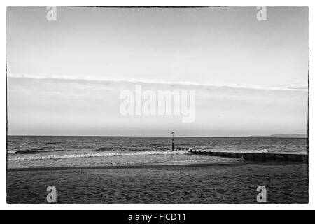 A black and white image of sunset light of a pier looking towards the sea at the beach with sand  and one strip of clouds in the Stock Photo