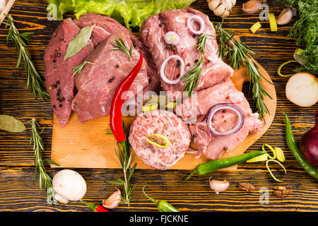 Various types of red raw meat with peppers, rosemary and spices on cutting board surrounded by other vegetables Stock Photo