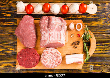 Top down view of matching pieces of raw red meat and pork on cutting board next to peppers, onion, rosemary sprig, tomatoes and Stock Photo