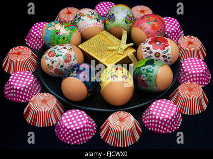 Easter eggs  with beautiful designs and a golden giftbox on black plate surrounded by cupcake holders Stock Photo