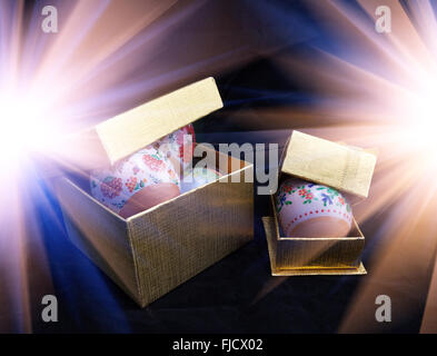 Ornate Easter eggs placed in golden gift boxes on dark background with light flares Stock Photo