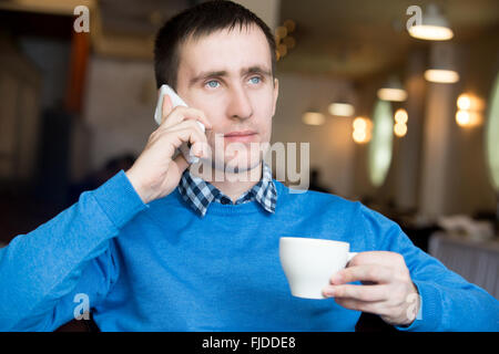 Portrait of casual relaxed smiling young business man sitting indoors in modern cafe, holding cellphone, using smartphone Stock Photo
