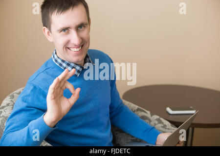 Portrait of casual attractive happy young man posing indoors, sitting in armchair with laptop, gesturing ok sign Stock Photo