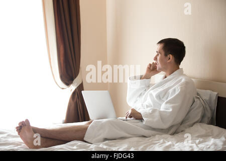 Portrait of attractive smiling young barefoot man wearing white bath robe sitting on bed with laptop, male model talking on cell Stock Photo