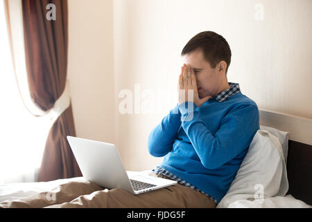 Portrait of young stressed man sitting on bed with laptop wearing smart casual clothing, holding his head in hands Stock Photo