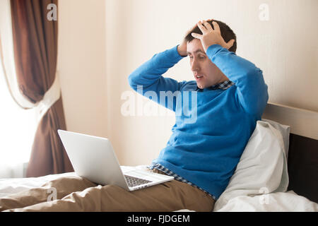 Portrait of young desperate man sitting on bed with laptop wearing smart casual clothing, holding his head in hands in dismay Stock Photo