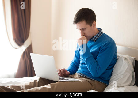 Portrait of young focused man sitting on bed with laptop wearing smart casual clothing, concentrated on work, male model Stock Photo