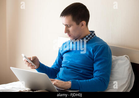 Portrait of attractive young man sitting on bed with laptop, using notebook and smart phone, making call, male model working Stock Photo