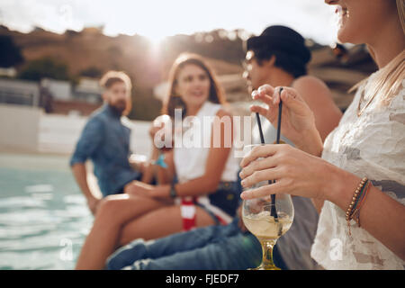 Woman holding a cocktail glass while sitting on the edge of swimming pool with friends. Young people enjoying a poolside party w Stock Photo
