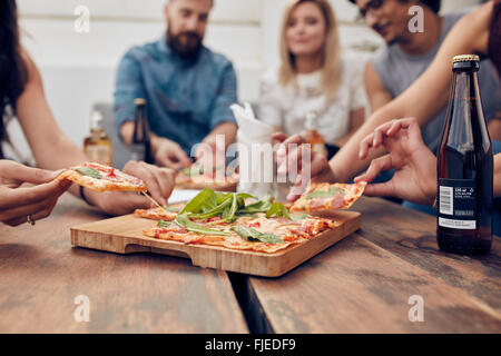 Close up shot of pizza on table, with group of young people sitting around and picking up a portion. Friends partying and eating Stock Photo