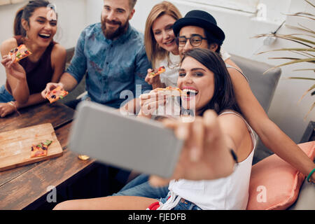 Group of multiracial young people taking a selfie while eating pizza. Young woman eating pizza her friends sitting around during Stock Photo