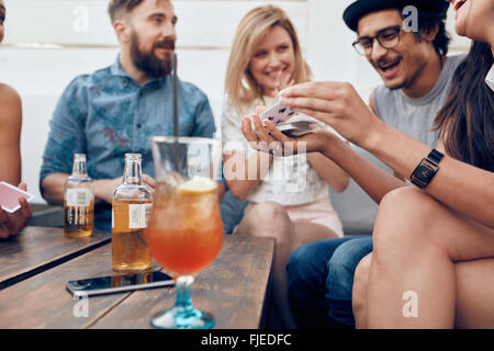 Group of young people sitting at a wooden table and playing cards. Woman shuffling cards while sitting with her friends in party Stock Photo