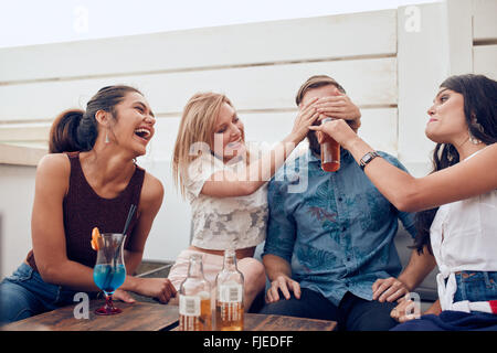 Shot of young friends sitting together enjoying party. Woman closing eyes of a man with another giving drink. Young people havin Stock Photo