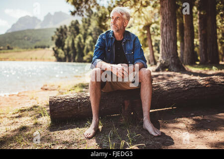 Portrait of a senior man sitting on a log near lake and looking away. Mature man sitting alone on lake. Stock Photo