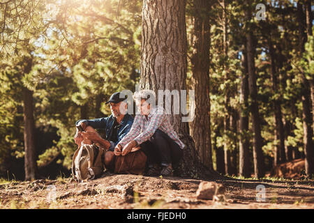 Portrait of mature couple sitting together under a tree with backpacks and compass. Senior man and woman in forest. Stock Photo